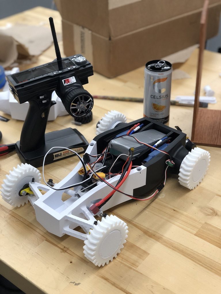 3D-printed remote-controlled race car with exposed drivetrain, battery, and transmitter alongside.